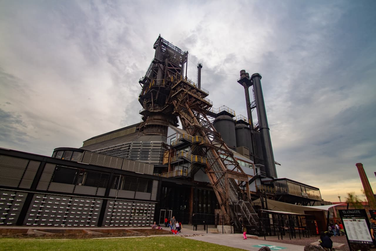 Una estructura industrial con varios niveles, compuesta por metal y cristal, donde se observa una torre metálica alta y tubos galvanizados. Alrededor hay personas y un espacio verde, bajo un cielo nublado.