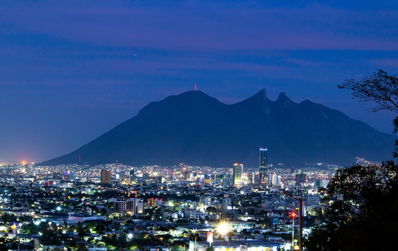 Vista nocturna de una ciudad iluminada, con un majestuoso cerro en el fondo. La montaña se destaca con un pico prominente y una antena en su cima, mientras que la urbe presenta rascacielos y una variedad de luces que crean un ambiente vibrante. El cielo tiene tonalidades azules y moradas, sugiriendo el ocaso.