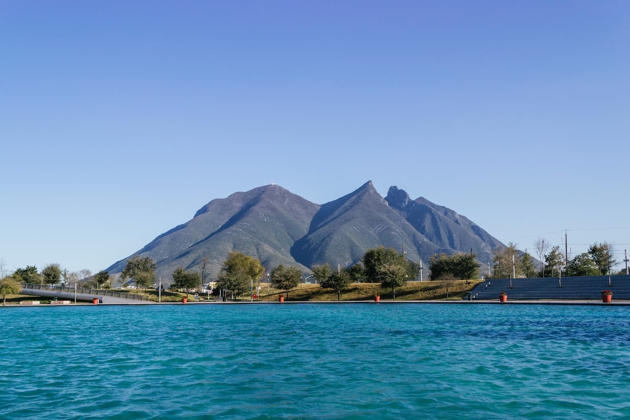 Se aprecia un paisaje con montañas en el fondo, un cielo despejado y azul, y un cuerpo de agua en primer plano que refleja su color turquesa. A la orilla, hay áreas verdes y árboles, así como una estructura de concreto que parece ser un pequeño puente.