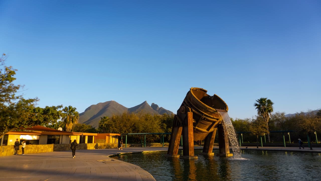 Un parque con una escultura metálica en forma de un recipiente volcado que vierte agua en una fuente. Al fondo se observan montañas y hay árboles y palmeras alrededor. Algunas personas caminando por el área. El cielo está despejado y el ambiente es sereno.