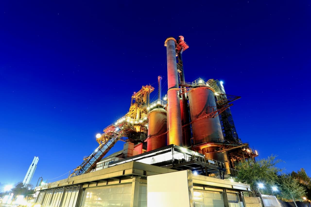 Estructura industrial vista de noche, con silos y tubos galvanizados iluminados en tonos cálidos, bajo un cielo azul profundo. En primer plano, se observa un edificio moderno y iluminado.