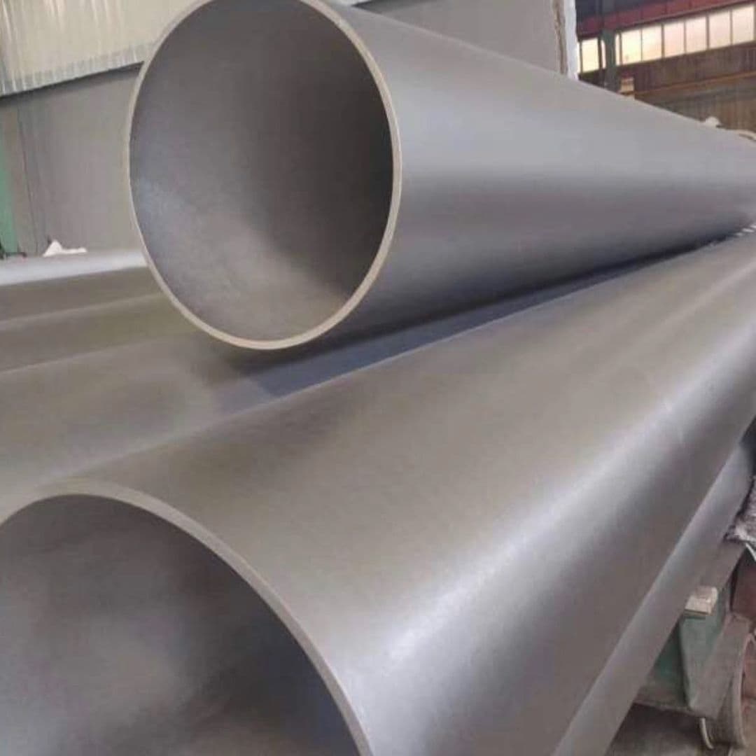 Se observan varios tubos galvanizados apilados, con un acabado liso y color gris. Algunos tubos están en posición horizontal y uno de ellos está en enfoque, mostrando su apertura desde un lado. El fondo parece ser un taller industrial.