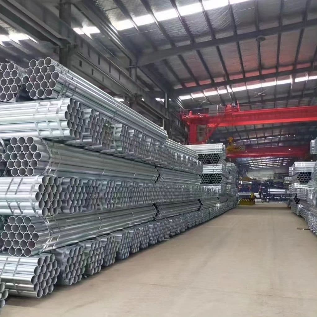 Interior de un almacén con varias filas de Tubería Galvanizada apiladas ordenadamente. La iluminación es brillante y hay maquinaria visible al fondo.